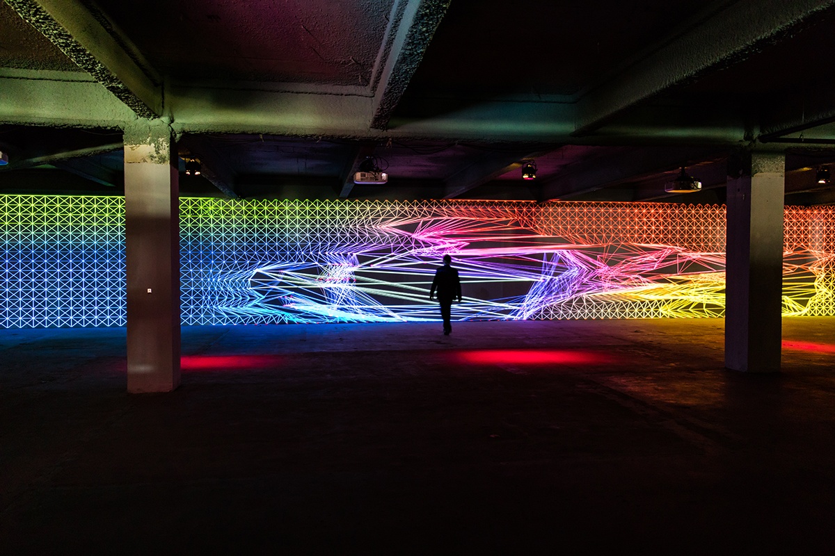 a person stands in a large room and looks at a light installation being projected on the far wall