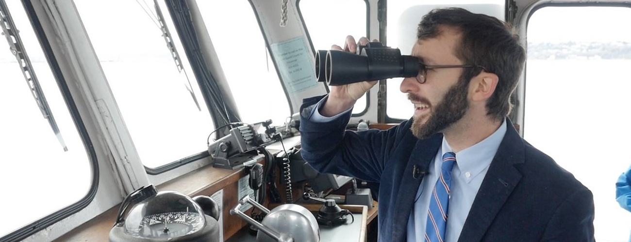 Boaz Frankel standing on the bridge of a boat, looking through binoculars off to the left of the image