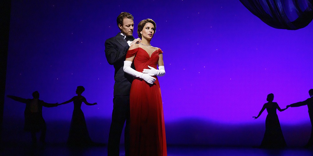 An actor and actress, both in formal wear, stand in the spotlight on stage.