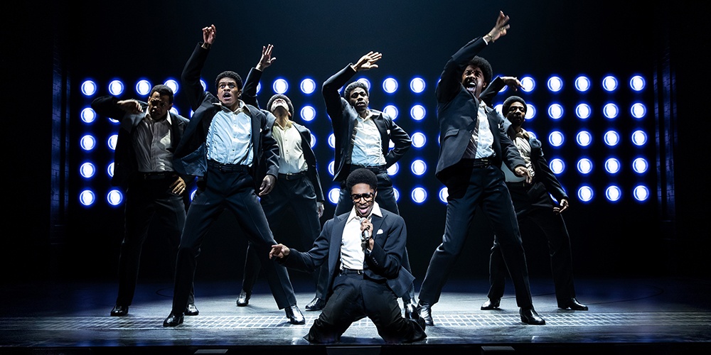 Ephraim Sykes, center, singing into a microphone while on his knees. Six other cast members dance behind him.