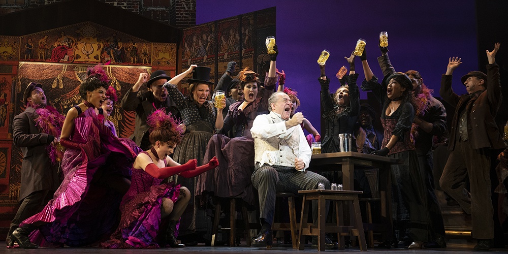 Adam Grupper, seated center, sings while surrounded by many castmembers who are also singing and raising their glasses.