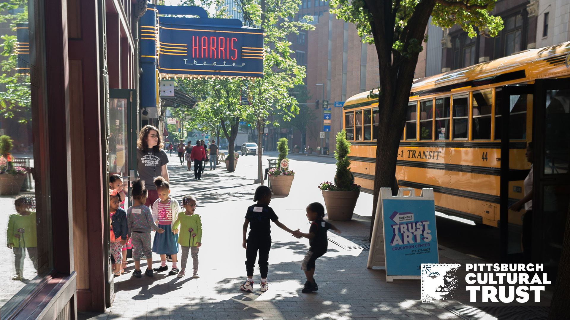 a zoom background of liberty avenue in front of the trustarts education center. a school bus with kids unloading is out front.