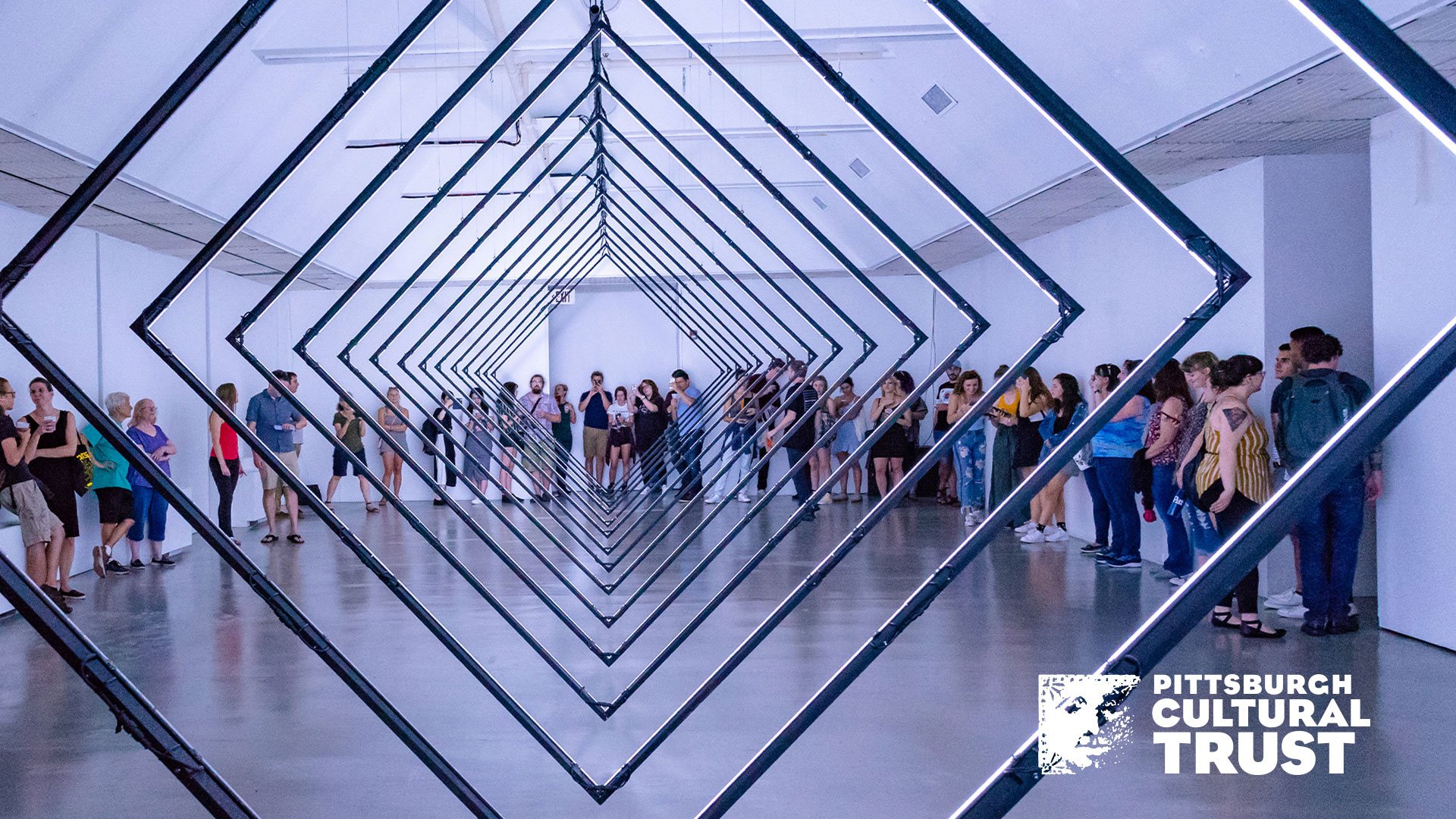 a zoom background of wood street galleries. a crowd of people watch a diamond-shaped light installation in the center of the room.