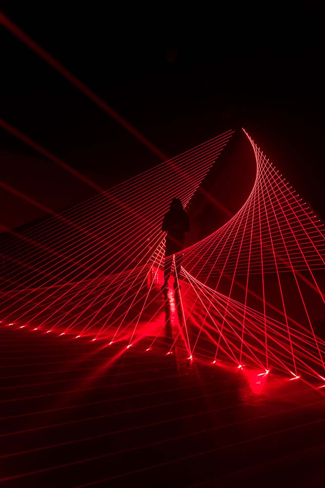  A detail view of Double Gemini. A silhouette of a person stands among red lasers that crisscross in front of and behind them