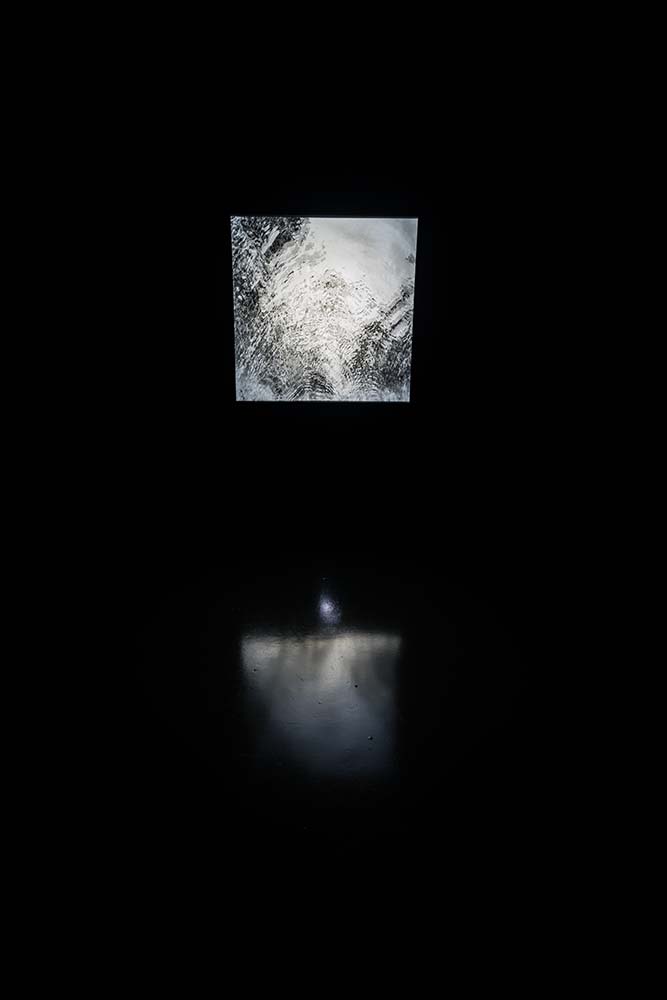 A wide view of Summoner, a square, strobing video projection in a dark room. An abstract black and white ripple pattern is seen on screen