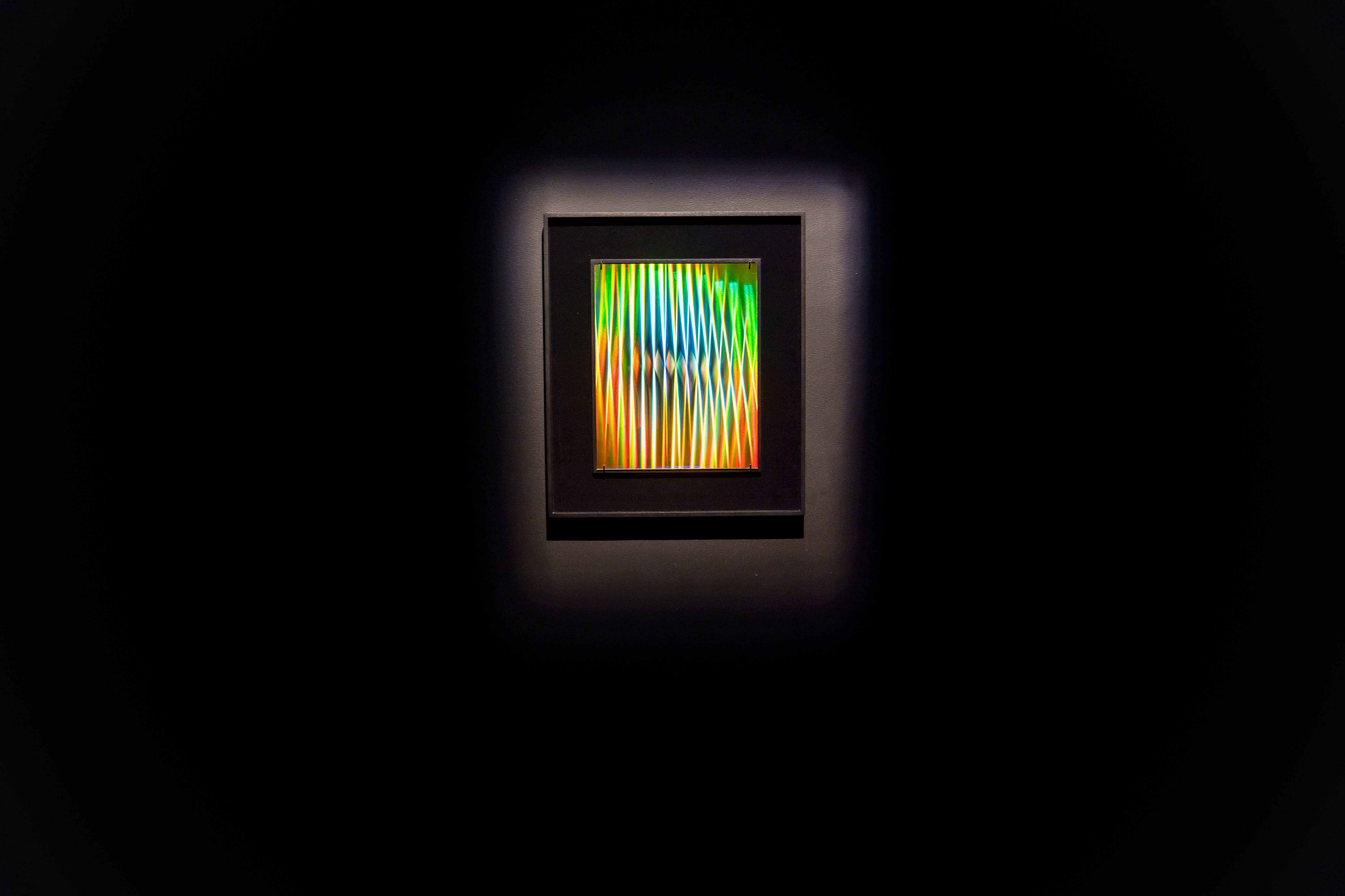 A piece from Gift Shop. A rainbow striped hologram in a black frame, hung on a black wall