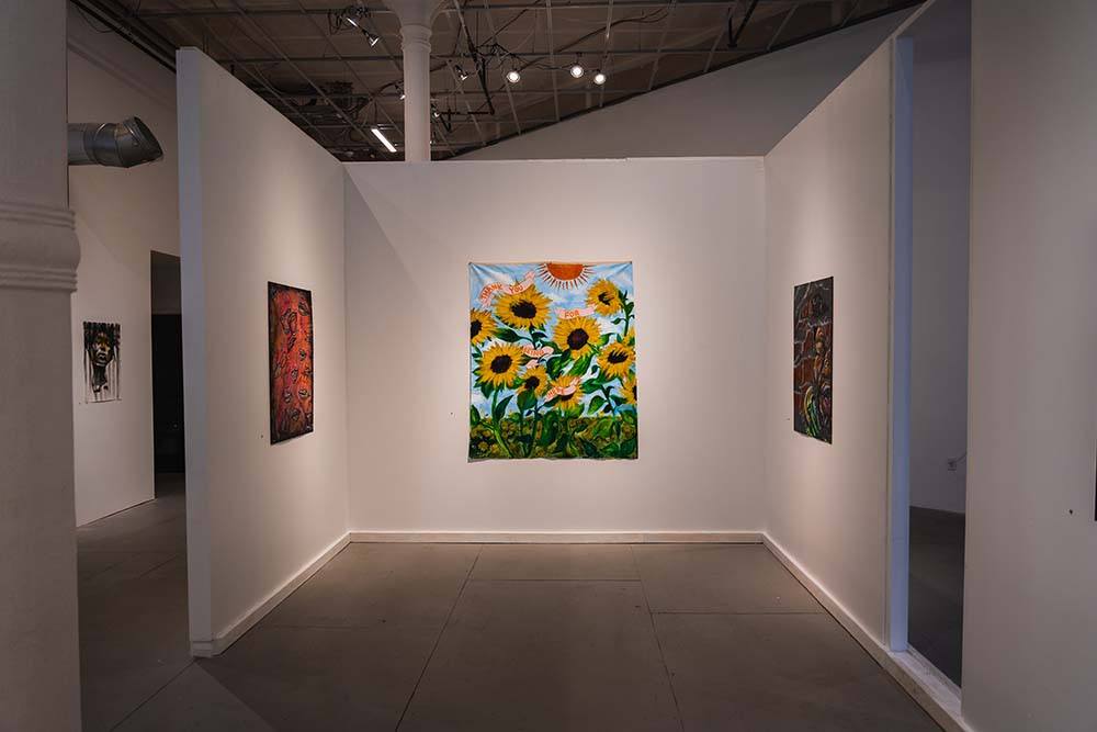 A painted wall hang depicting sunflowers and blue sky. Two abstract paintings hang on perpendicular walls to the right and left