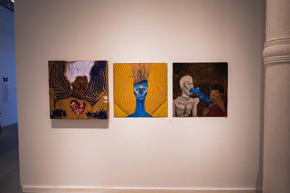Three square paintings hung in a row. The first is a portrait of a man, the second is a blue-skinned humanoid with the top of its head exploding, and the third is a monster sucking the life out of a human