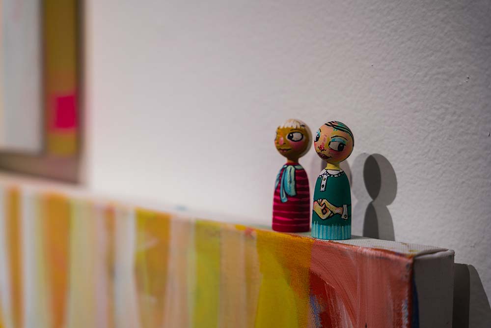 A detail of two small painted wooden figurines sitting on the top edge of a wall-hung canvas