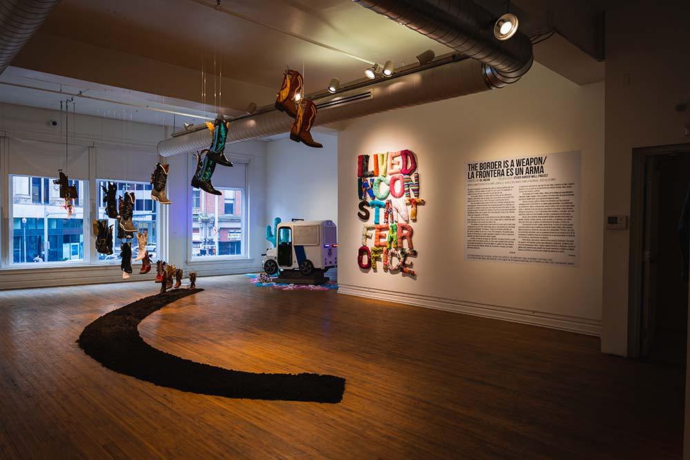 A wide view of the front portion of the exhibit, with the gallery statement on the left, a fabric piece hanging in the center, and hanging boots on the left