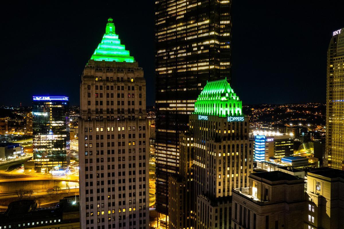 Night view of city skyscrapers with two prominent buildings featuring bright green illuminated rooftops, surrounded by other lit-up buildings and city lights in the background.
