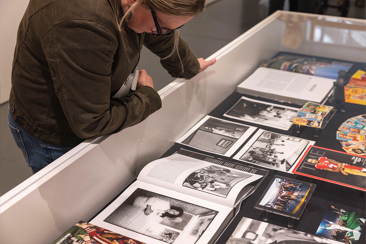A person wearing glasses and a brown jacket leans over a display case, closely examining open books and black-and-white photographs, along with magazines and colorful printed materials arranged inside.