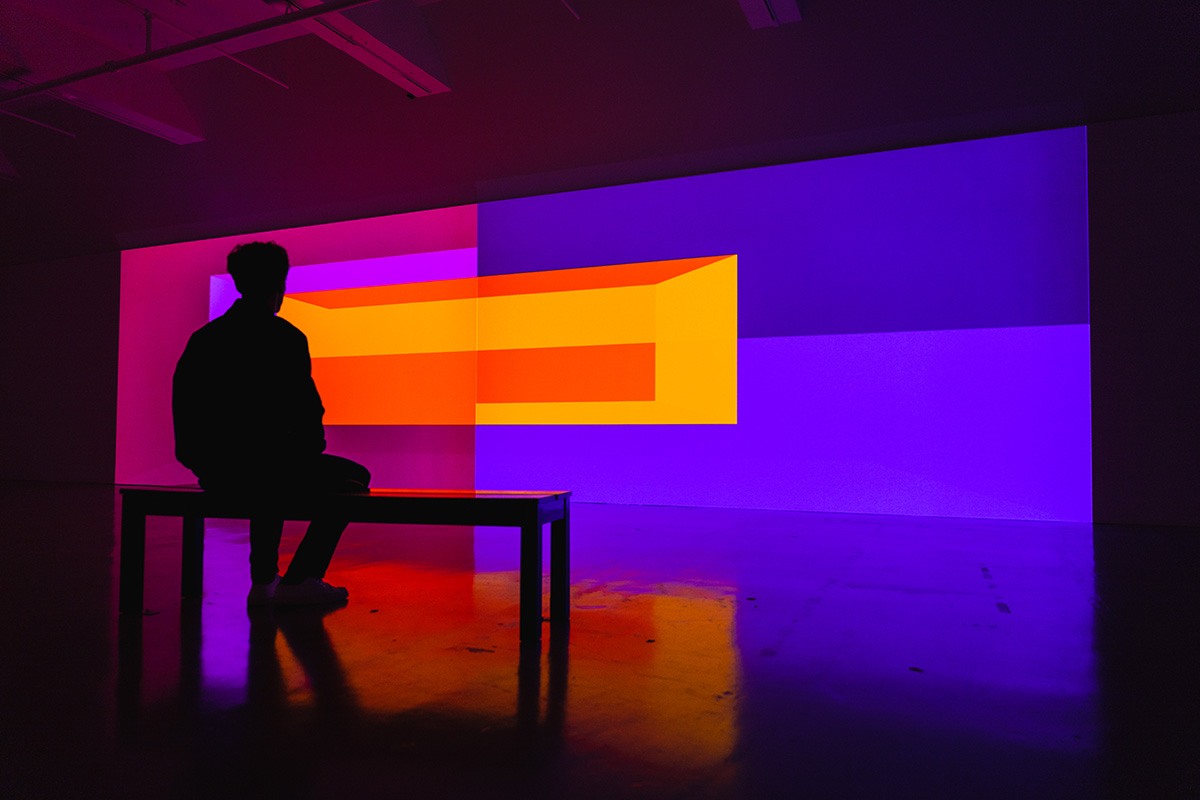 a long wall in a dim gallery space has a colorful geometric design projected on it. a person sits on a bench in front of the wall.