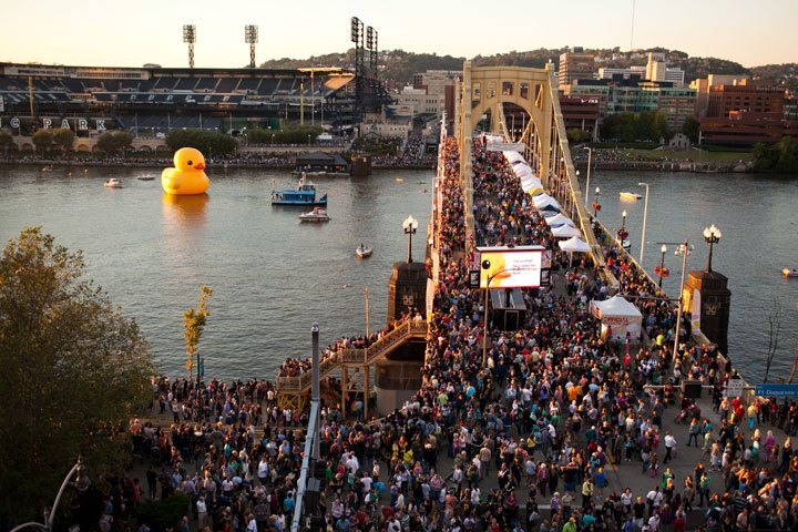 A large crowd stands on and around a yellow bridge. A large yellow rubber duck, the reason for all the people, floats in the water.