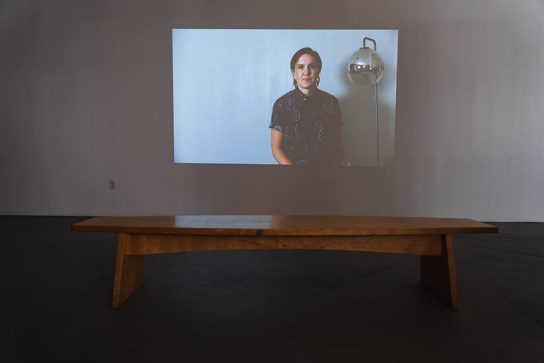 a projected video shows a person on screen looking at the camera. a wooden bench is in the foreground.