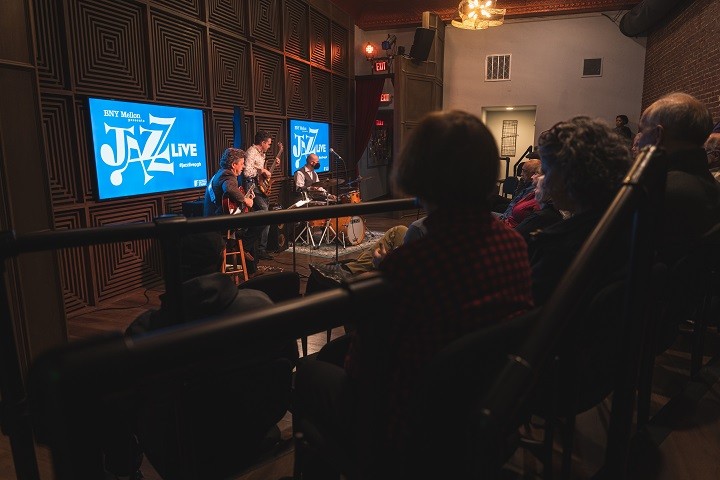 Two guitar players and one drummer perform in front of a seated audience.