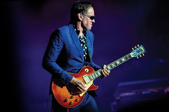A man in a blue suit and dark shades plays an electric guitar.