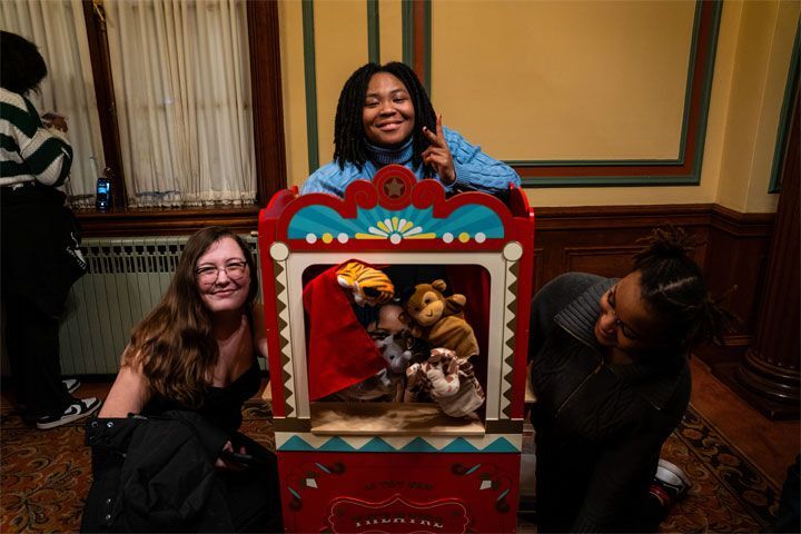 Three students crouch around a kid's puppet theater and smile as one student pops their head through the opening.