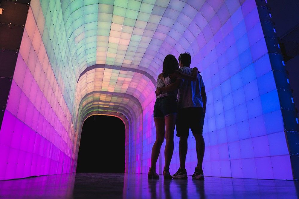 a couple standing arm in arm in front of wide, arched tunnel made of multicolored light up blocks