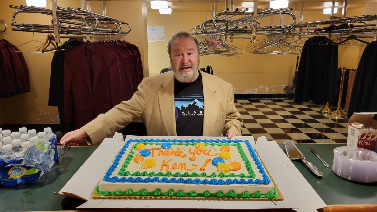 a man in a brown sport coat and black t-shirt stands in from of a large cake that says 'thank you ken'