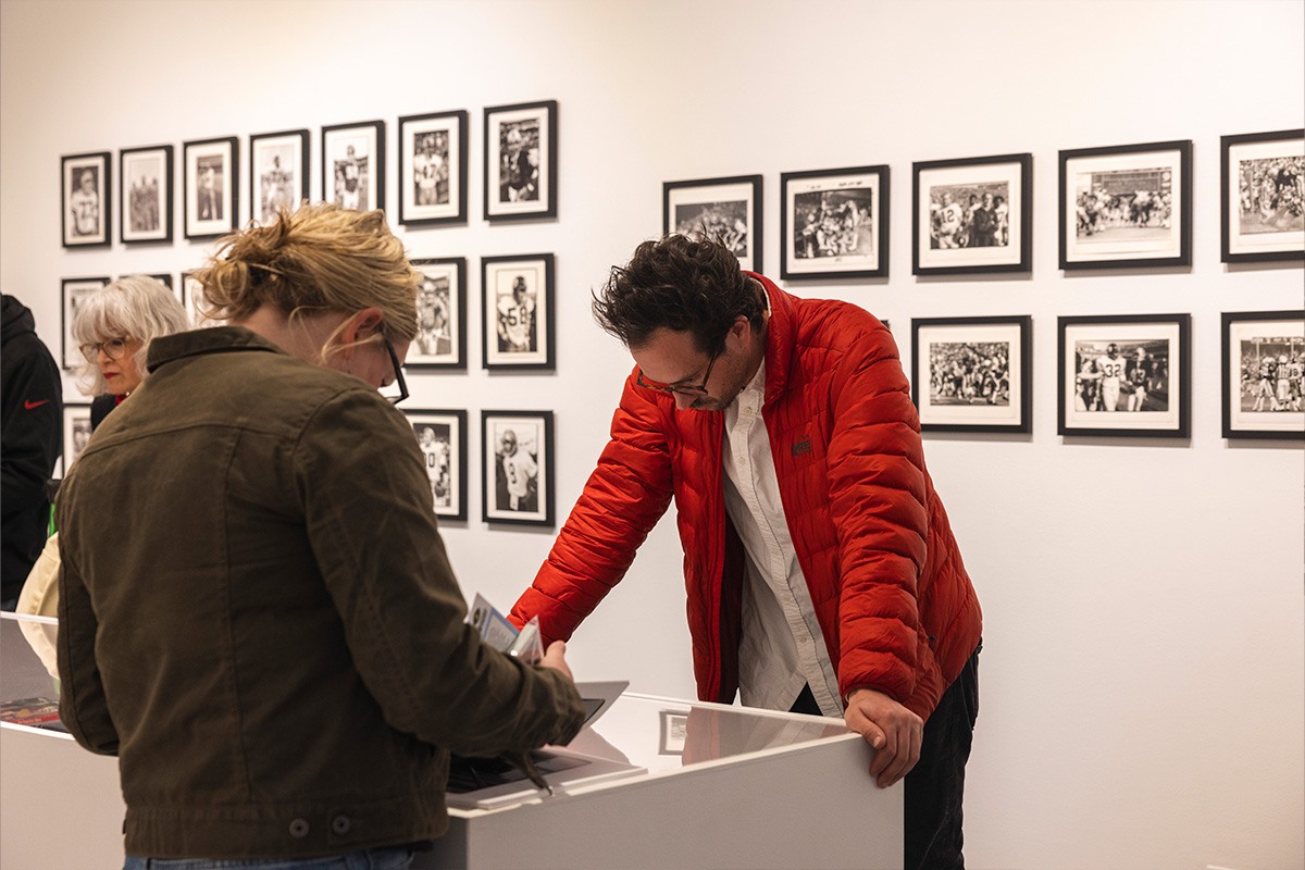 Two people view and discuss items in a gallery with numerous black-and-white framed photos on the wall. One person wears a red jacket, and the other has blonde hair and glasses, looking at objects on a display table.