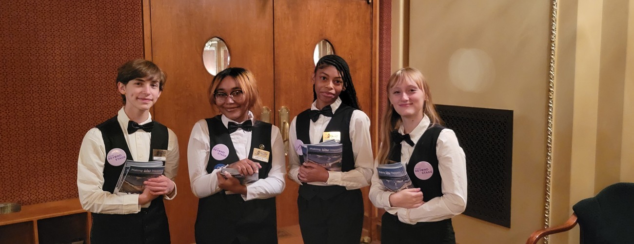 Four high students pose in their volunteer uniforms, a white button-down shirt, a black vest, and a black bow tie, in the Benedum Center lobby.