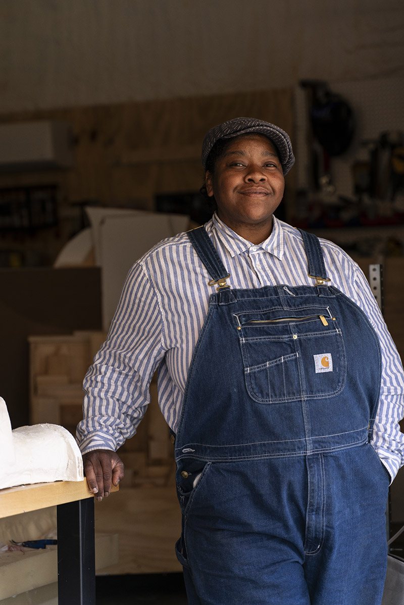 A Black woman in overalls and a grey hat.