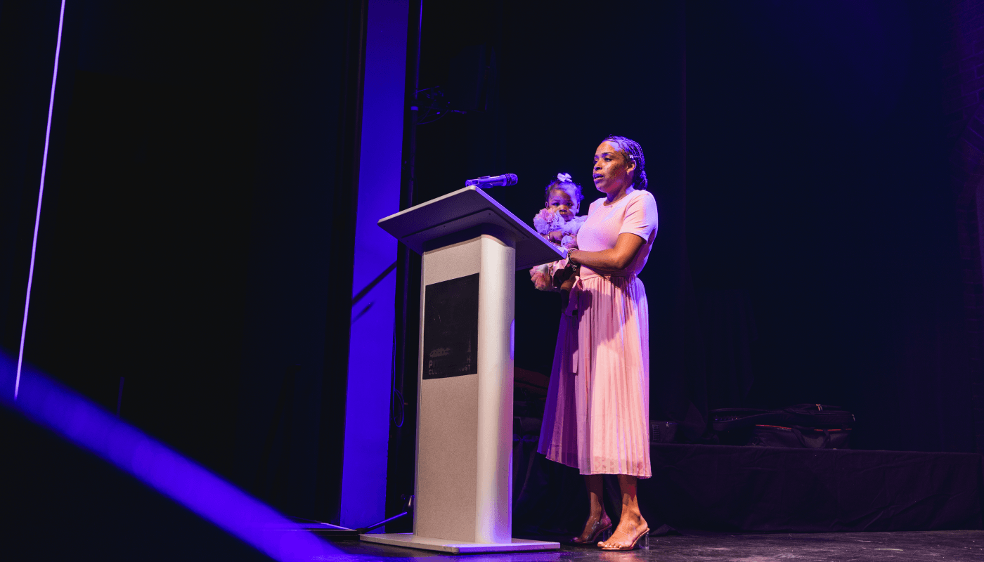 Black woman speaks at a podium while holding her baby.