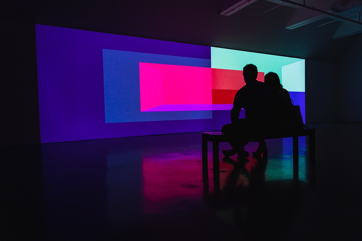 a long wall in a dim gallery space has a colorful geometric design projected on it. two people sit next to each other on a bench in the middle of the room.