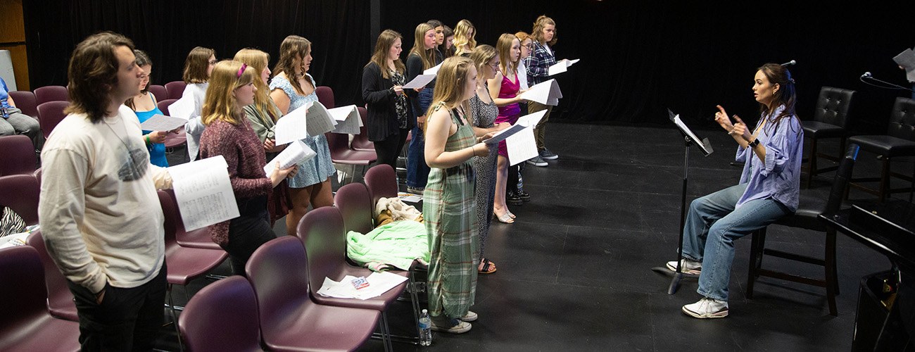 a group of young adults all hold sheet music as they stand in front of an instructor