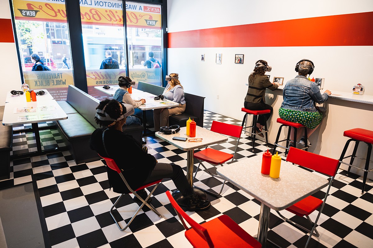 five people sit in a gallery space dressed to resemble a vintage diner. each person has a VR headset and headphones on