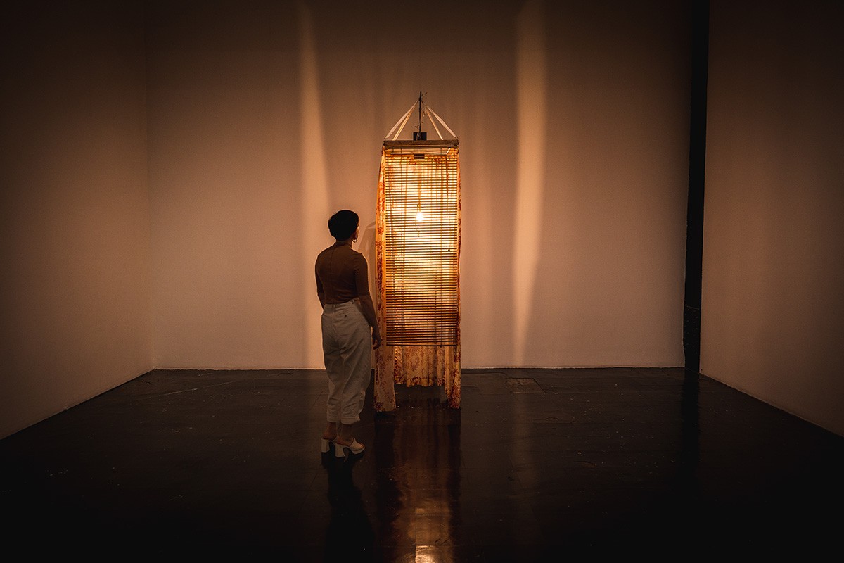 a dim gallery room with a tall, thin sculpture sitting in it. a single lightbulb in the center of the piece casts a yellow glow over the space. a person stands next to it.