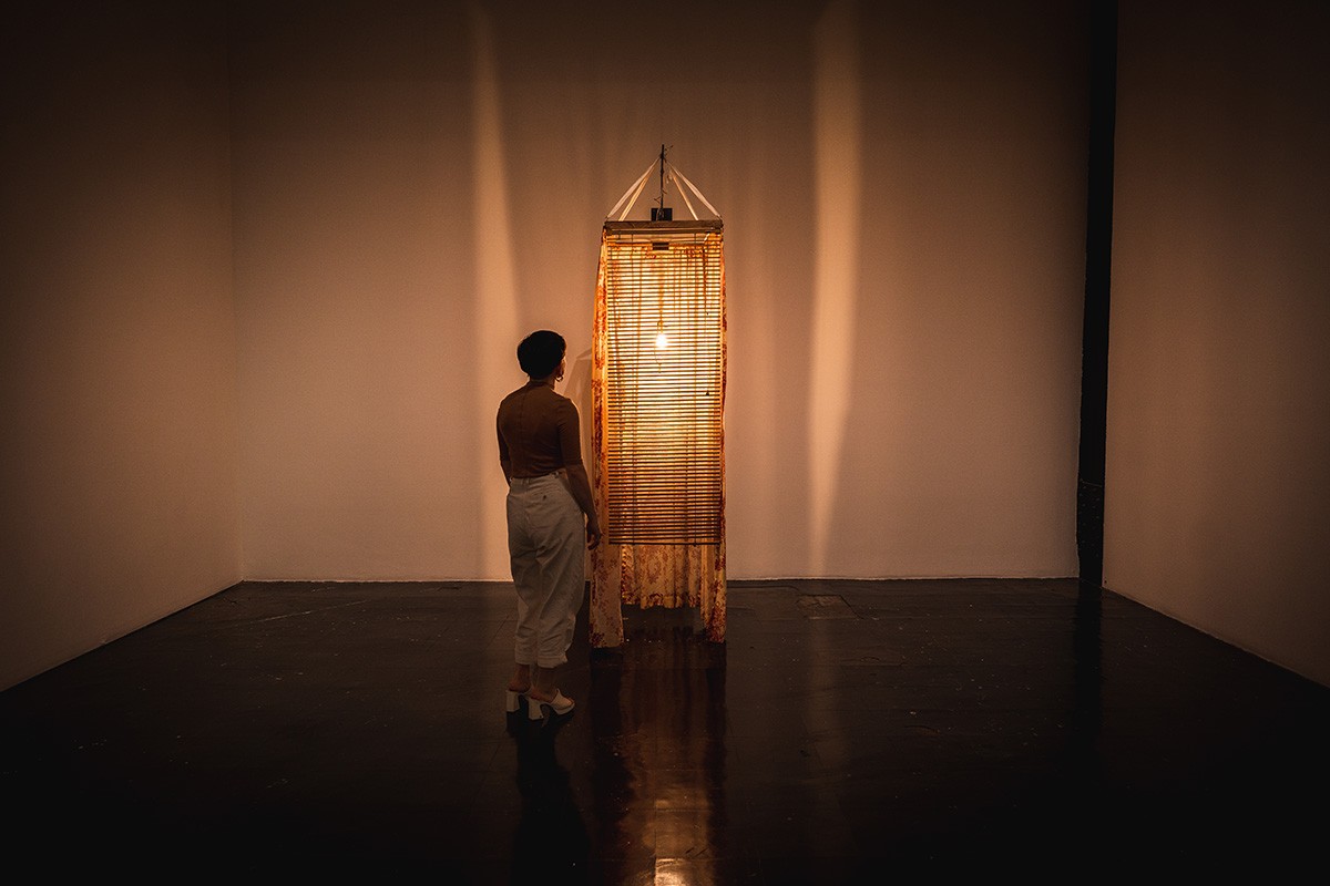 a dim gallery room with a tall, thin sculpture sitting in it. a single lightbulb in the center of the piece casts a yellow glow over the space. a person stands next to it.