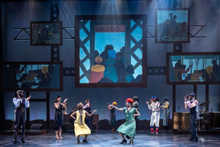 Black dancers in dressy costumes dance on stage with five works of art behind them.