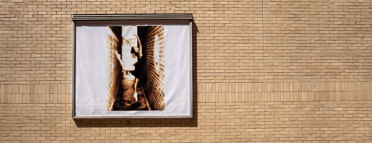 A framed sepia-toned photograph hangs on a beige brick wall. The photo shows a narrow alley with a person reaching toward a brick wall, creating a sense of depth and contrast against the plain background.