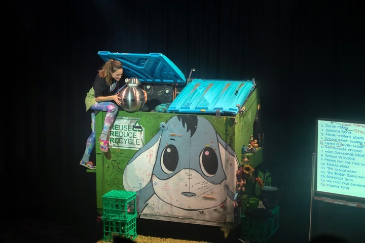 A woman on stage climbs up a dumpster that has a cartoon animal on the front. She's holding a small, round, metal robot.