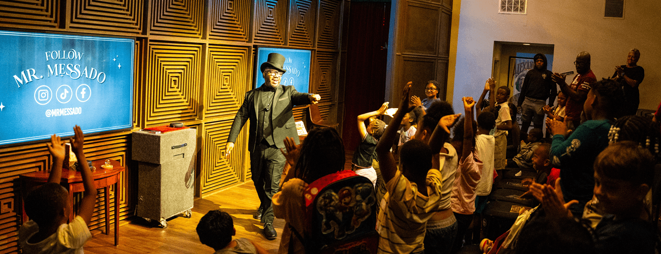 A man in his magician's uniform: black top hat, black vest, and a black suit, smiles while pointing at a group of kids with their hands raised.