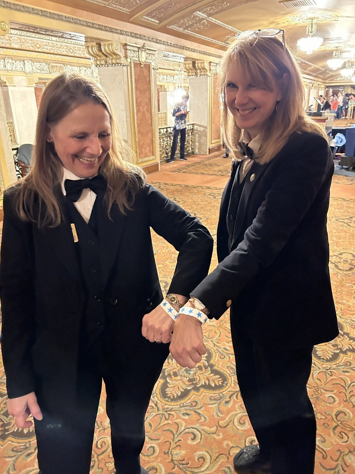 two white women in black suits smile and hold out wristbands they are wearing