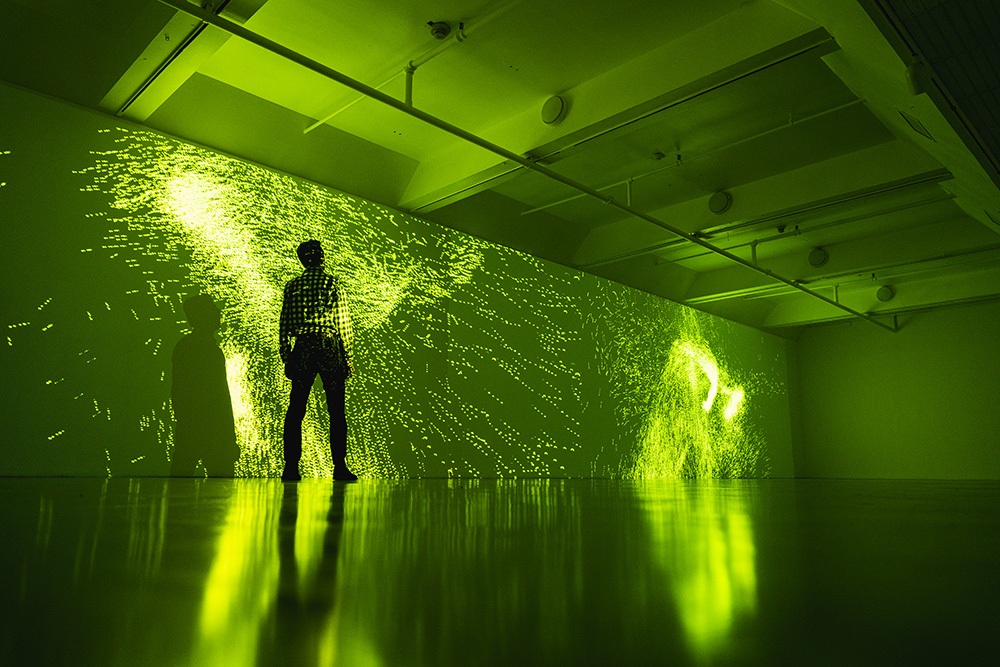 a long wall in a dim gallery space has splatter patterns of green light projected on it. a person stands in front of one splatter.