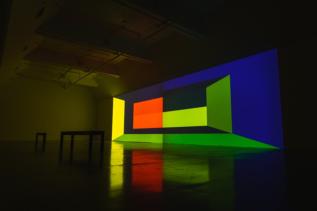 a long wall in a dim gallery space has a colorful geometric design projected on it. two benches sit in the middle of the room.