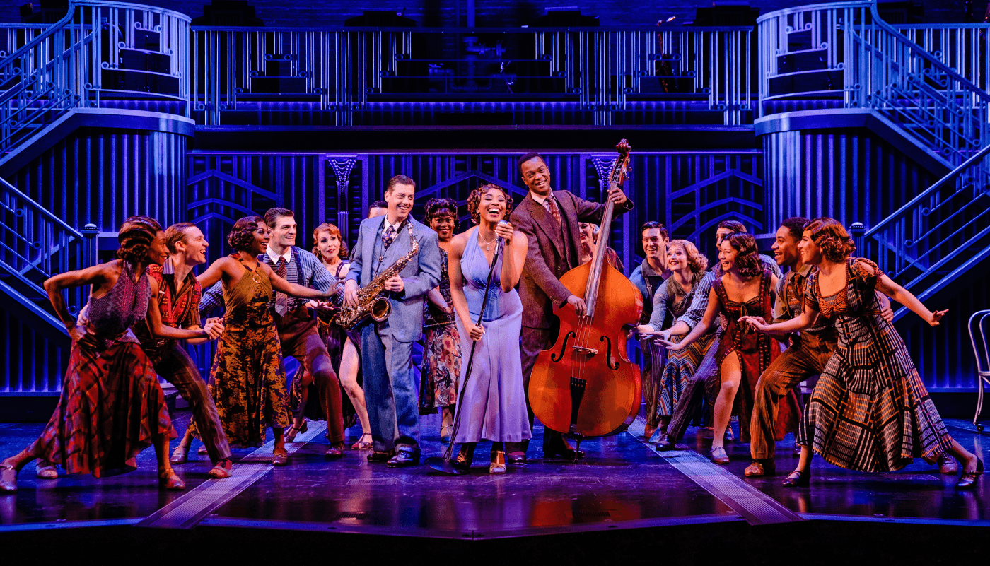 A group of musicians and singers on stage. One musician holds an upright bass while a woman sings into a microphone in the middle of the group.