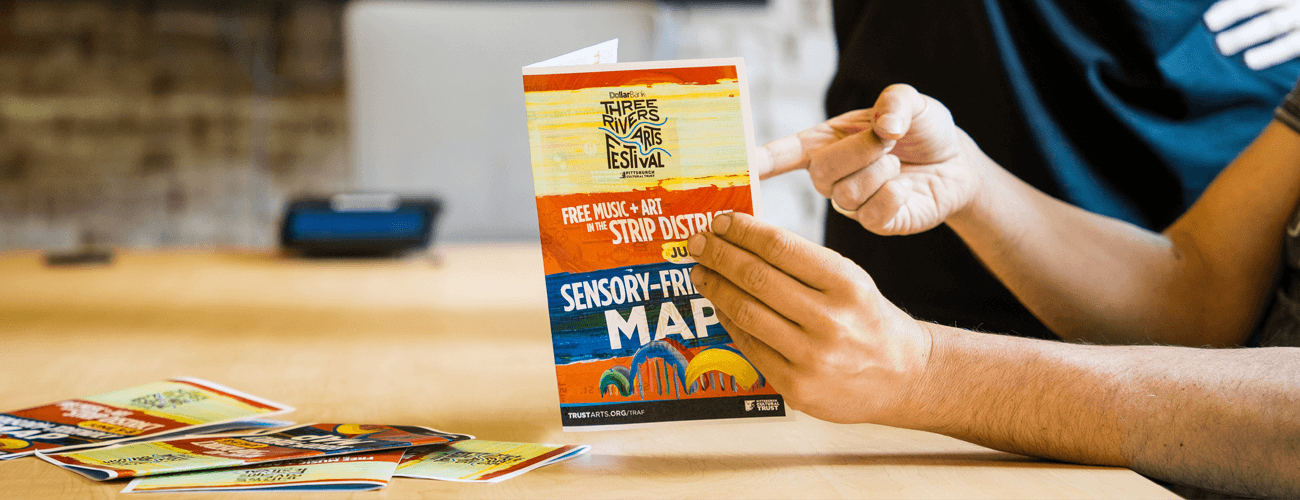 A person holds and points to a colorful event brochure that reads Free Music + Art in the Strip District and Sensory-Friendly Map; other brochures are spread on a table.