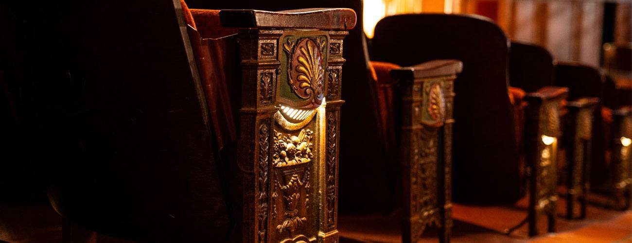  A close up of the side of a Benedum Center seat. The wooden side panel on the side of the seat is ornately carved and a light glows from the middle.