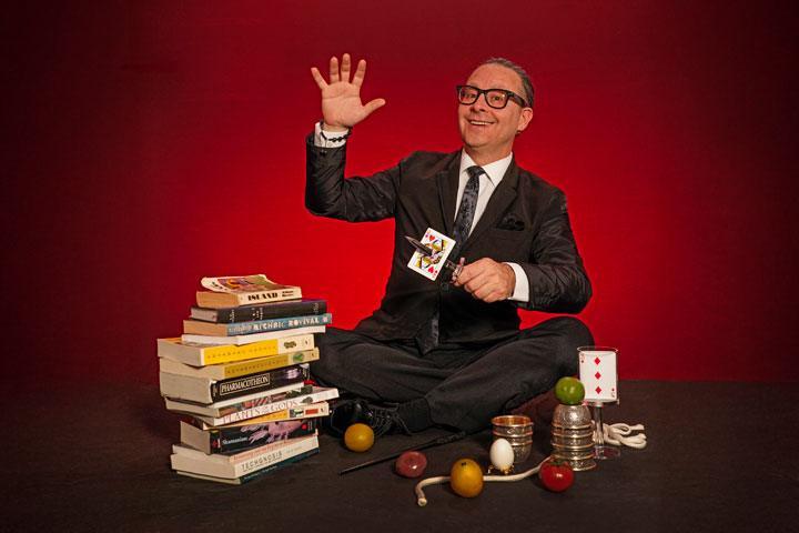A man in a suit and black tie sits cross legged, holding up one hand in the air and the other, he's pierced a knife through a card. He sits around a pile of books, a rope, a cup and ball trick, and a card.