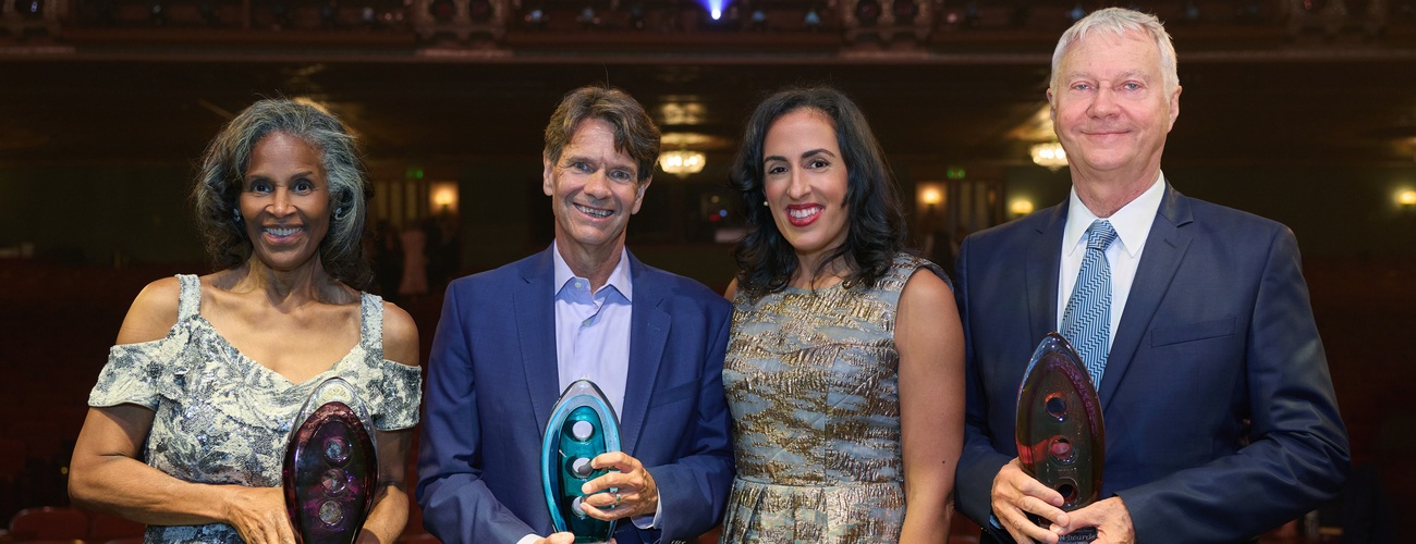 Four people dressed formally stand side by side on a stage, smiling and holding glass awards, with a blurred theater audience in the background.