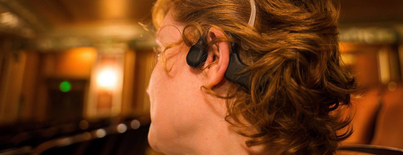A woman sits in a theater seat wearing glasses, a hearing aid, and bone conduction headphones.