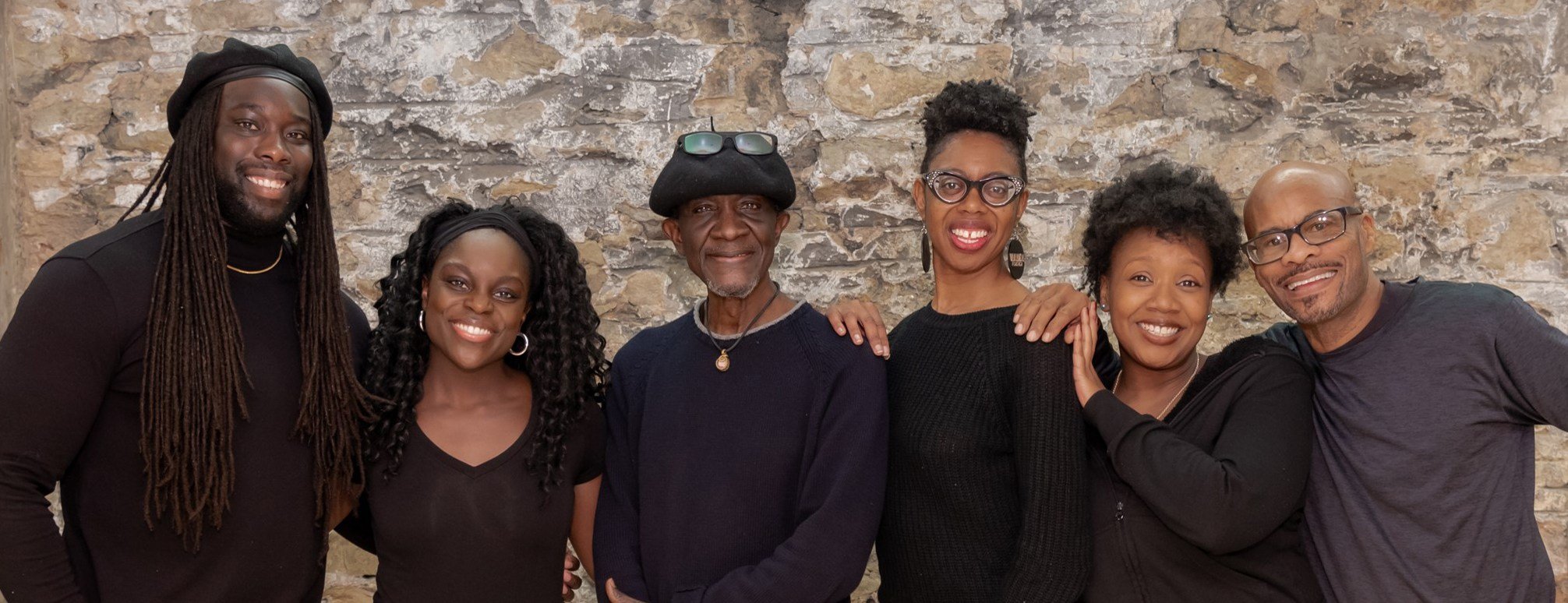 a group six black teaching artists wearing dark toned clothing stand together and smile