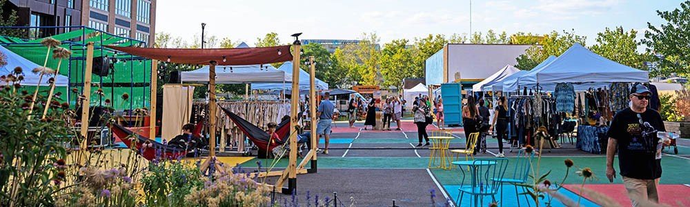 Outdoor market with various vendor tents, plants, and colorful flooring. People are walking, shopping, and some are sitting in hammocks.