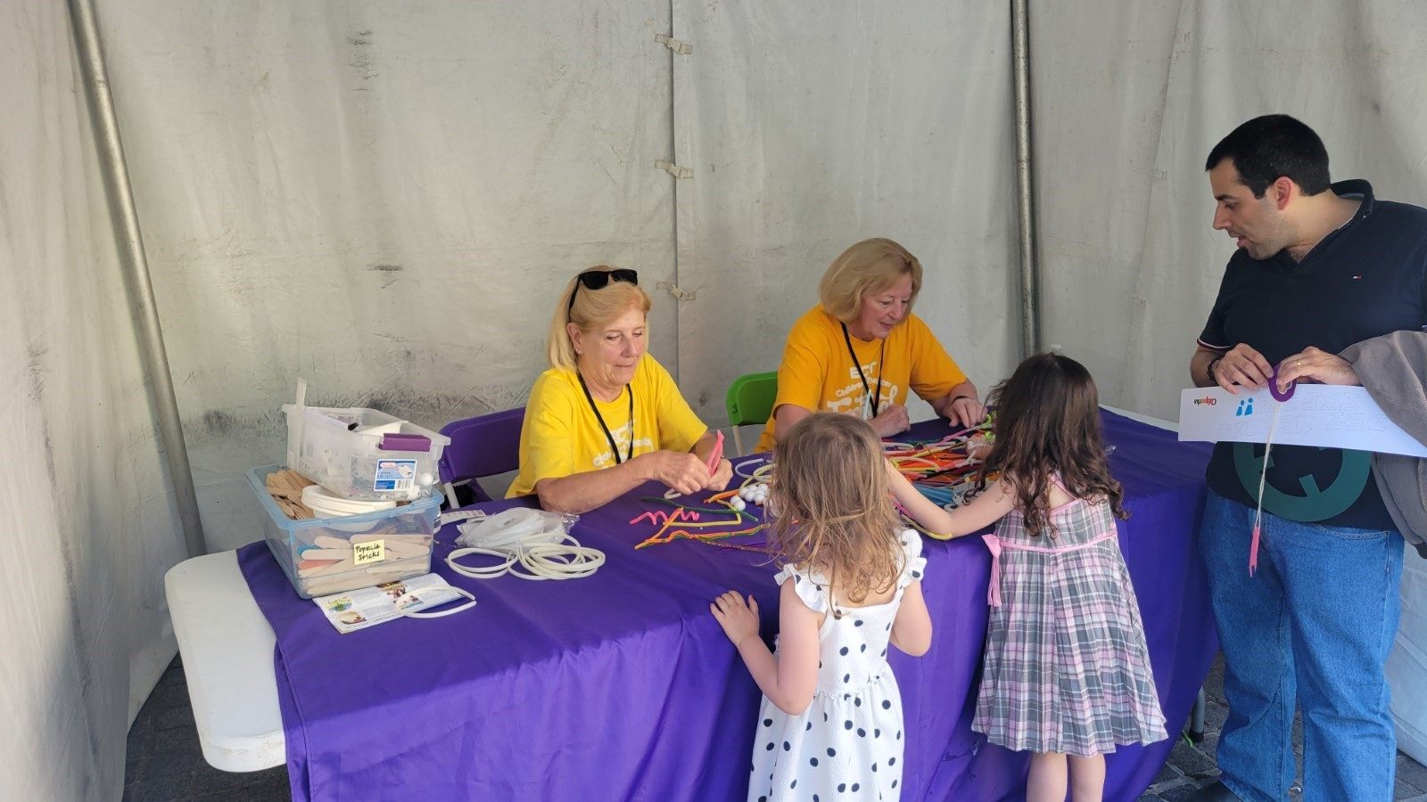 Two volunteers in yellow t-shirts sit at a table with colorful crafts. They are interacting with two young children.