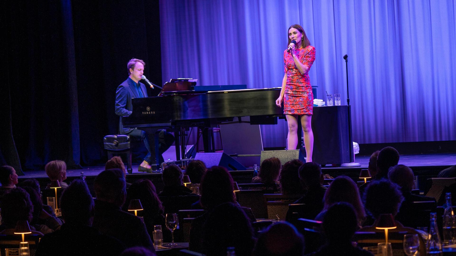 A woman in a red dress sings into a microphone on stage next to a man playing a grand piano, with an audience seated at tables watching the performance under purple lighting.